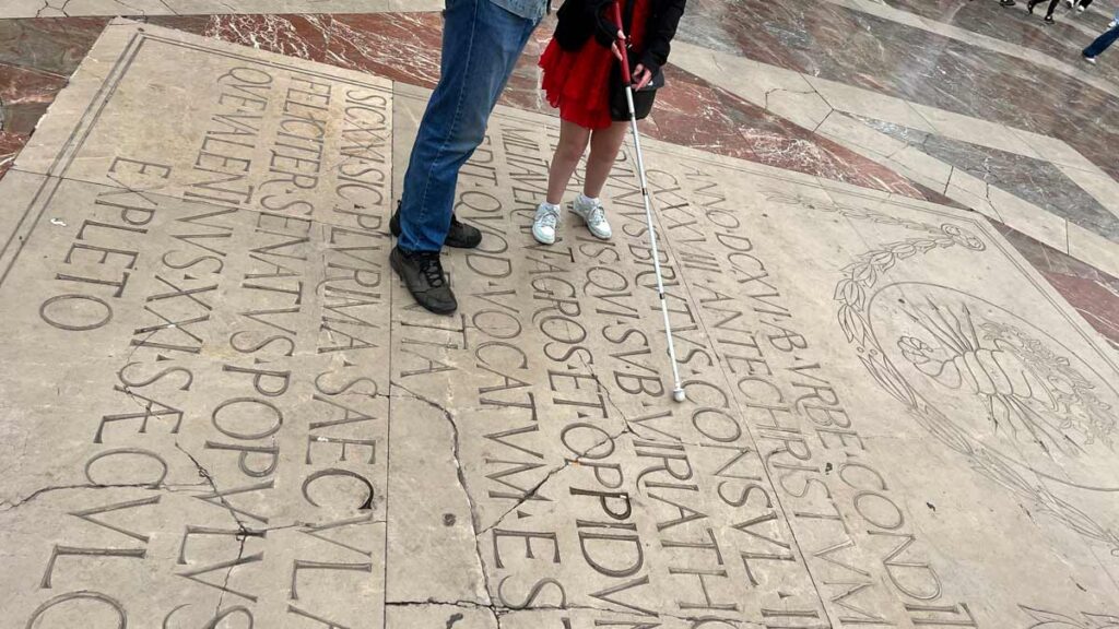 A photo of a large plaque on the floor, with Latin writing on it. You can see the bottom half of Sassy, wearing a red dress, black jacket and white trainers, next to a man wearing blue jeans and black shoes. She is holding the white cane over the Latin inscription.