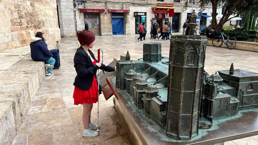 Sassy wearing a red dress, black jacket and white shoes, holding her white cane in one hand, and touching a giant replica of the Cathedral in Valencia with the other.