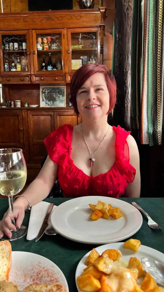Sassy sitting down at a table, smiling at the camera. She is wearing a red dress and a necklace with a red stone. She is holding a glass of white wine and has a plate with patatas bravas in front of her. Behind her you can see an old fashioned wooden cabinet with drinks, glasses, and old photos inside.