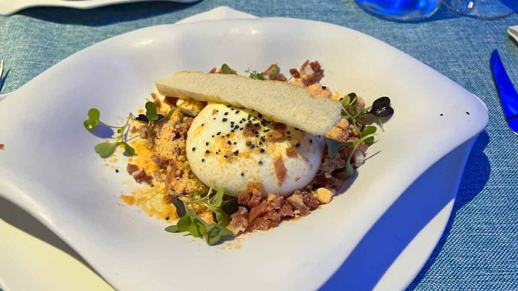 A plate of buratta cheese over Spanish jamon and crumbs, with a slice of crispy bread on top. The dish is served inside a deep plate, with the shape of a sting ray.