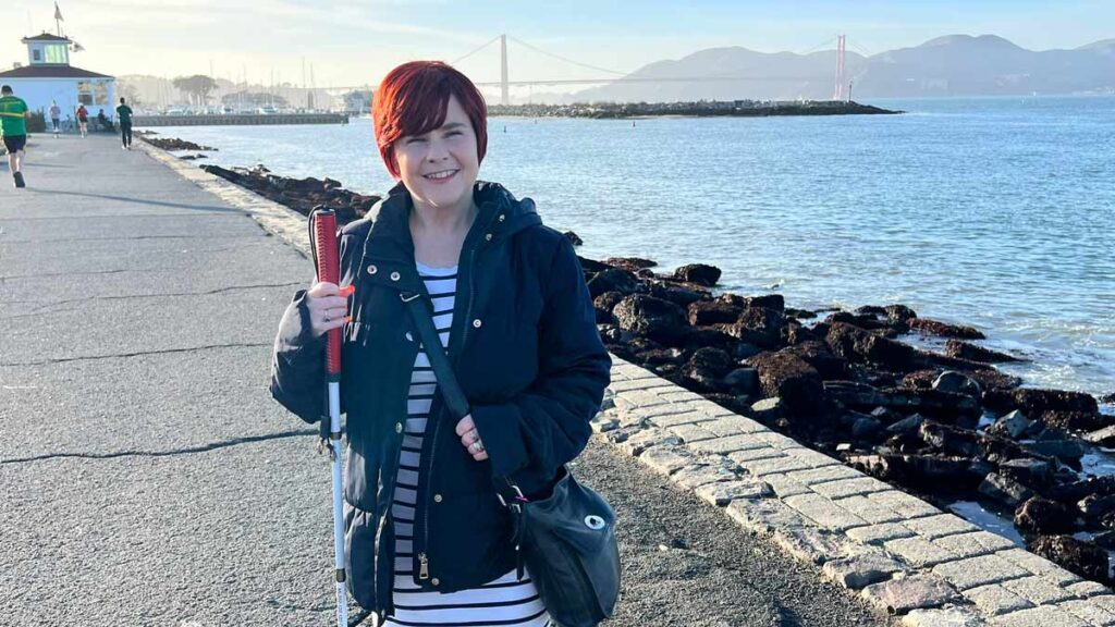 Sassy smiling at the camera, with Golden Bridge behind her. She is wearing a black and white stripe dress, and a black jacket on top. She is holding her white cane with a red handle in one hand, and her the other one on her black crossbody bag. The light behind her is very warm. 