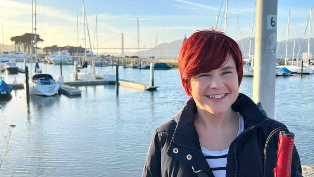 A close-up of Sassy smiling at the camera. She has short red hair, a black jacket and a black and white stripe tshirt. Behind her you can see a harbour with boats and yachts anchored on docks. In the background you can spot the golden bridge in a warm light.
