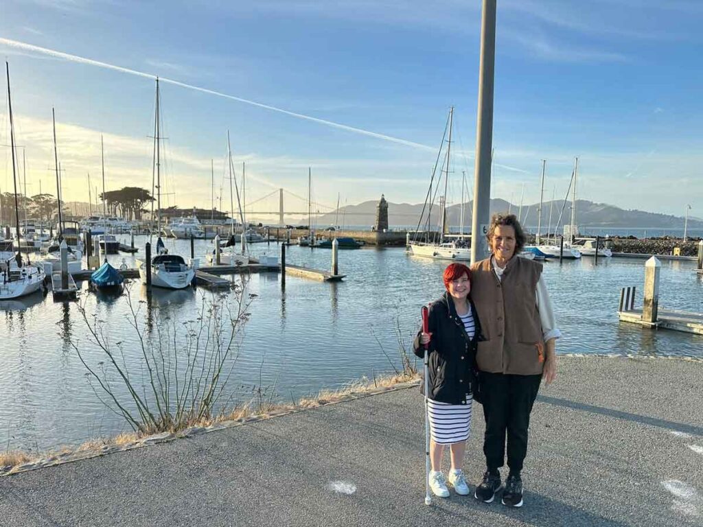 Sassy and Connie posing together next to a harbour, with the Golden Gate bridge behind them. Sassy is wearing a black and white stripes over the knee dress, with a black jacket over, and white trainers. Connie is wearing a brown vest, white jumper, black trousers and black trainers. 